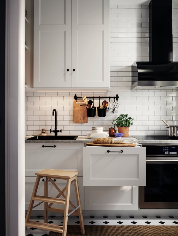 AXSTAD matte white kitchen fronts, a step stool, black handles, and a countertop with bread, plates, and potted plants.