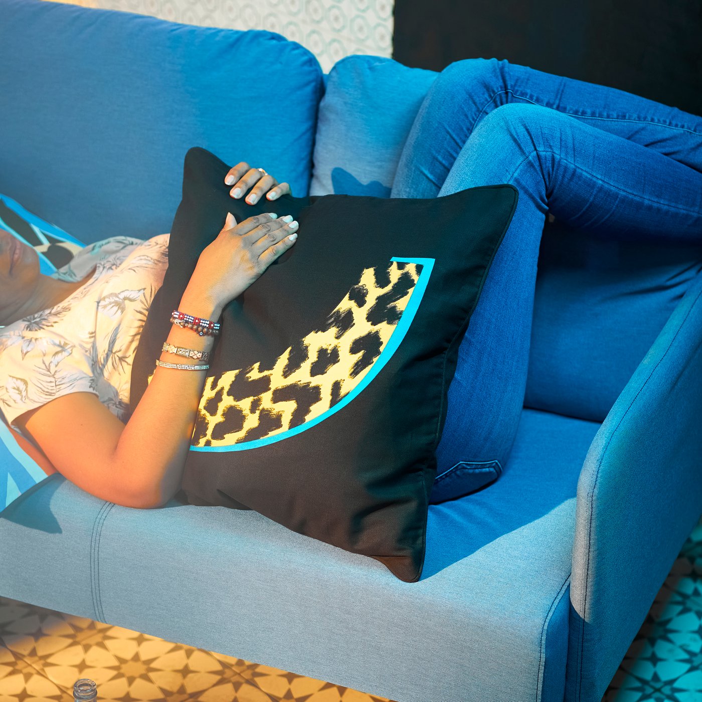A person in jeans is lying on a turquoise sofa, hugging a large pillow with a black/yellow ÖMSESIDIG cushion cover.