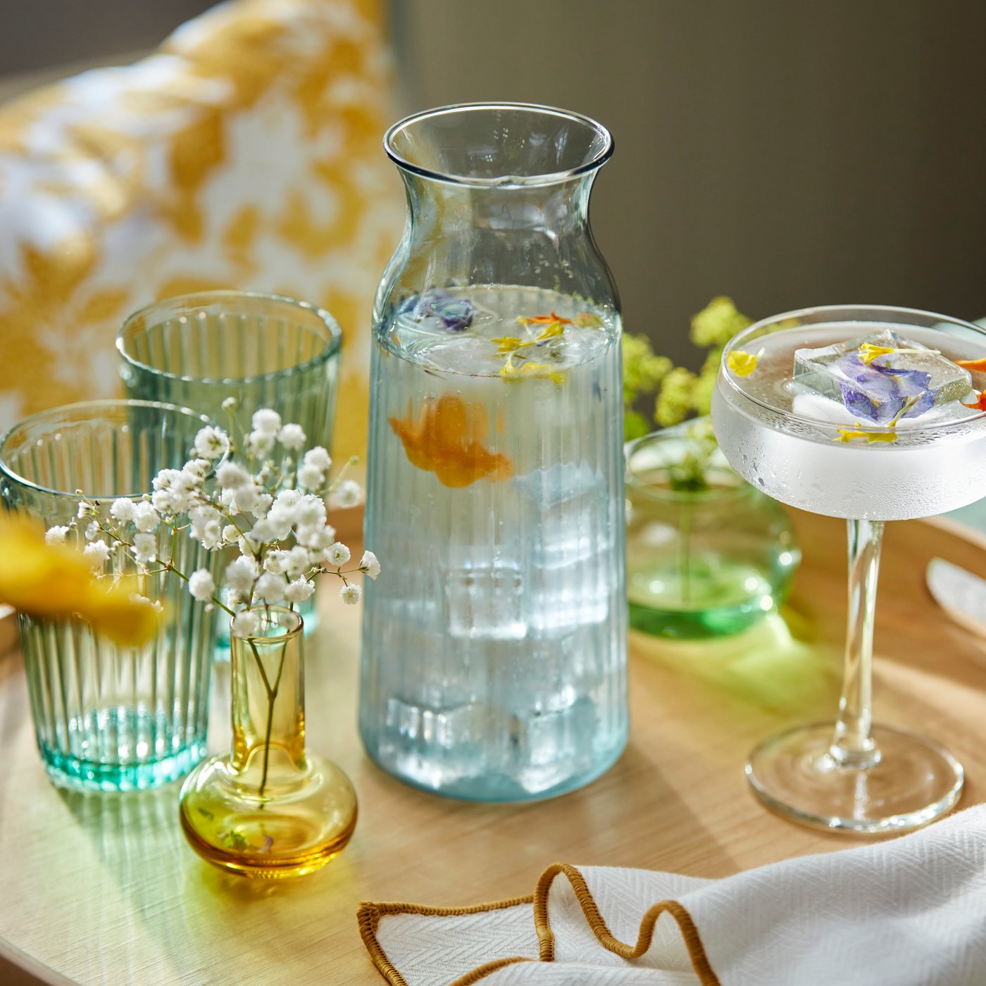 Glass carafe filled with water, surrounded by pastel drinking glasses and small floral decorations on a wooden table.