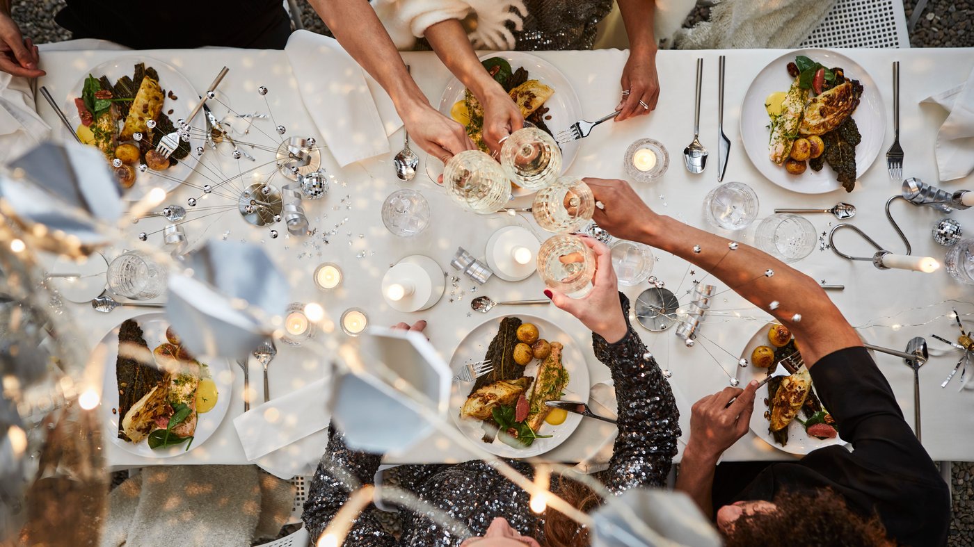 Bird’s-eye view of silver and white New Year’s Eve table setting, raising toast with FRÖJDA champagne coupes.