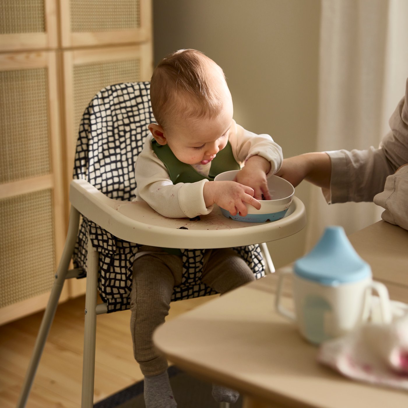 A baby sitting in the SANDKATT highchair playing with a BÖRJA plate/bowl next to an adult supervising.
