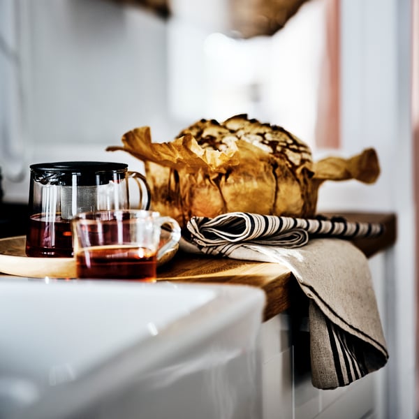 Un rangement de cuisine blanc avec un plan en bois, un mug IKEA 365+ et une théière RIKLIG remplie de thé dessus, à côté d’une gros gâteau.