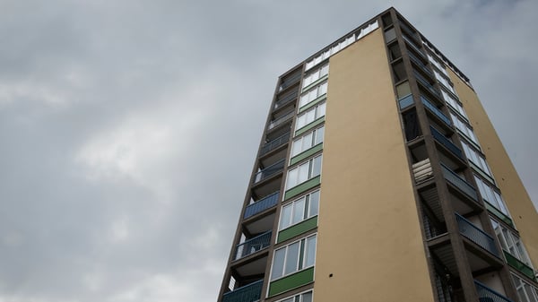 A beige and green high-rise apartment building with corner balconies and windows against a cloudy sky.