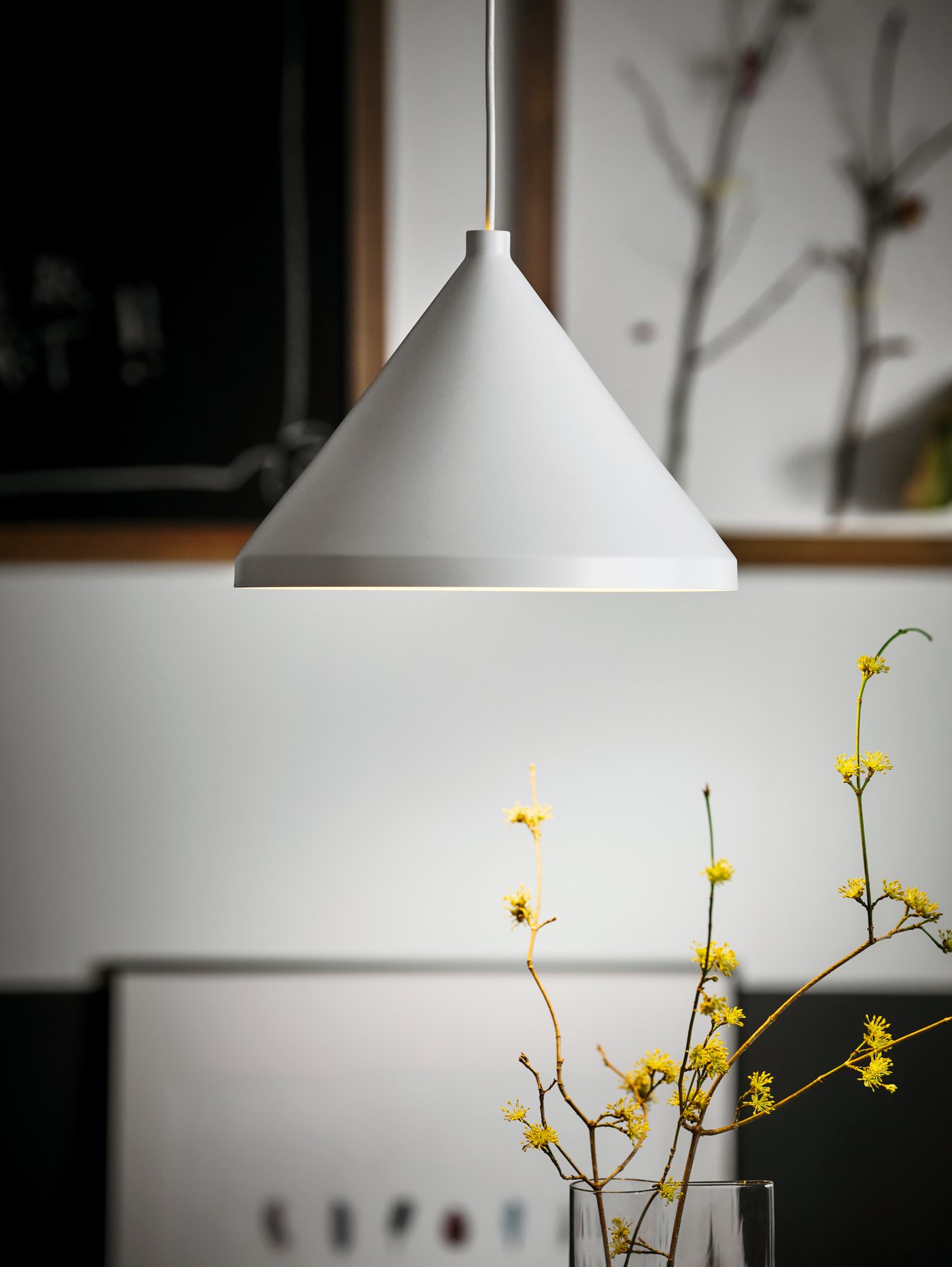Une suspension NÄVLINGE blanche au-dessus de brindilles aux fleurs jaunes dans un vase. Des œuvres encadrées sont accrochées à un mur à proximité.