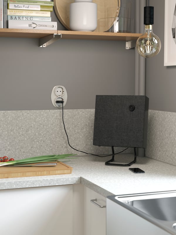 A small square shaped black speaker standing in the corner of a grey worktop next to a chopping board.