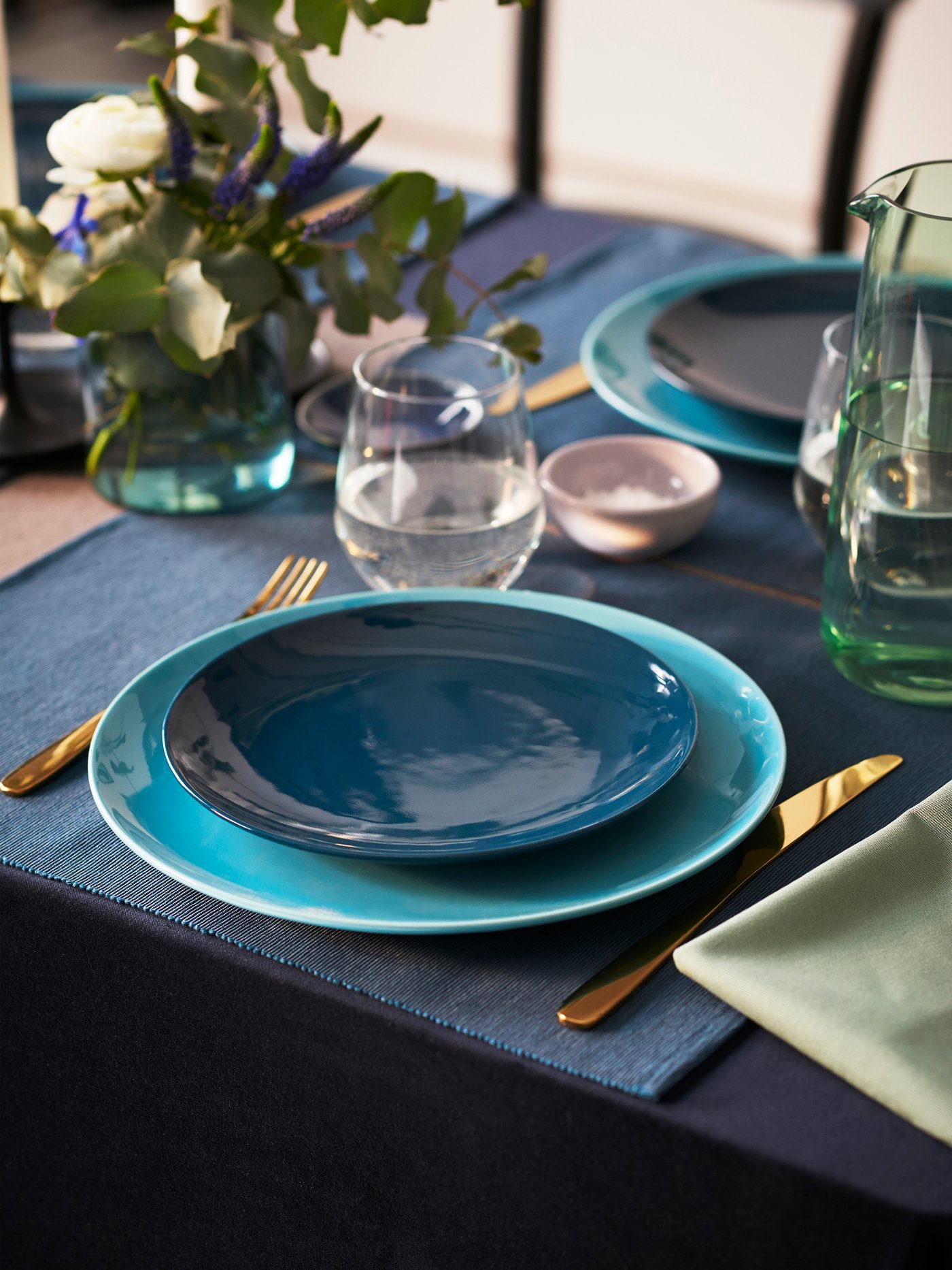 Un puesto en una mesa con individuales y platos FÄRGRIK azules, cubiertos TILLAGD dorados y, frente a los platos, un vaso transparente IVRIG. 