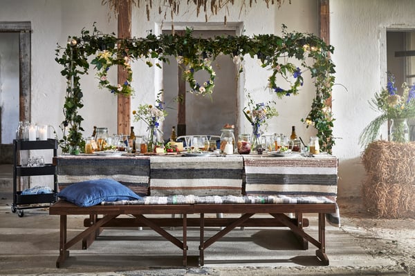 A long wooden table, festively decorated with garlands and floral wreaths. Food, drinks and a black IKEA RÅSKOG serving trolley are visible. A rustic atmosphere.