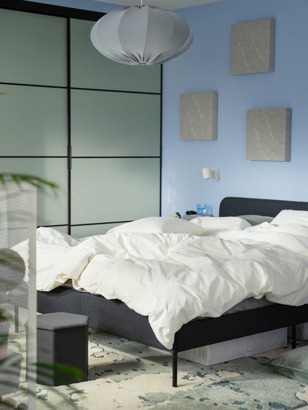 A dark grey SLATTUM upholstered bed, with a STUK storage case underneath it, stands on a green/off-white RINGKLOCKA rug.