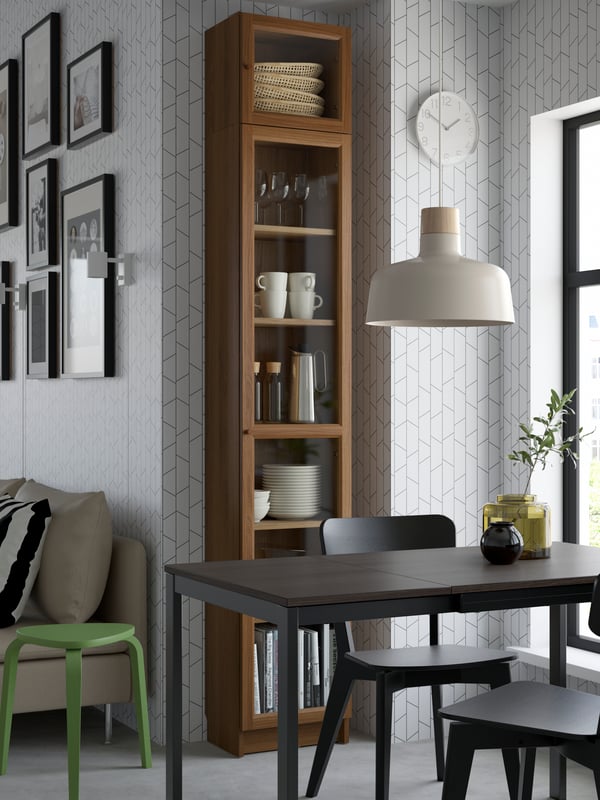 A BILLY/OXBERG bookcase with glass doors filled with tableware and glassware, standing in an odd corner in a dining area.