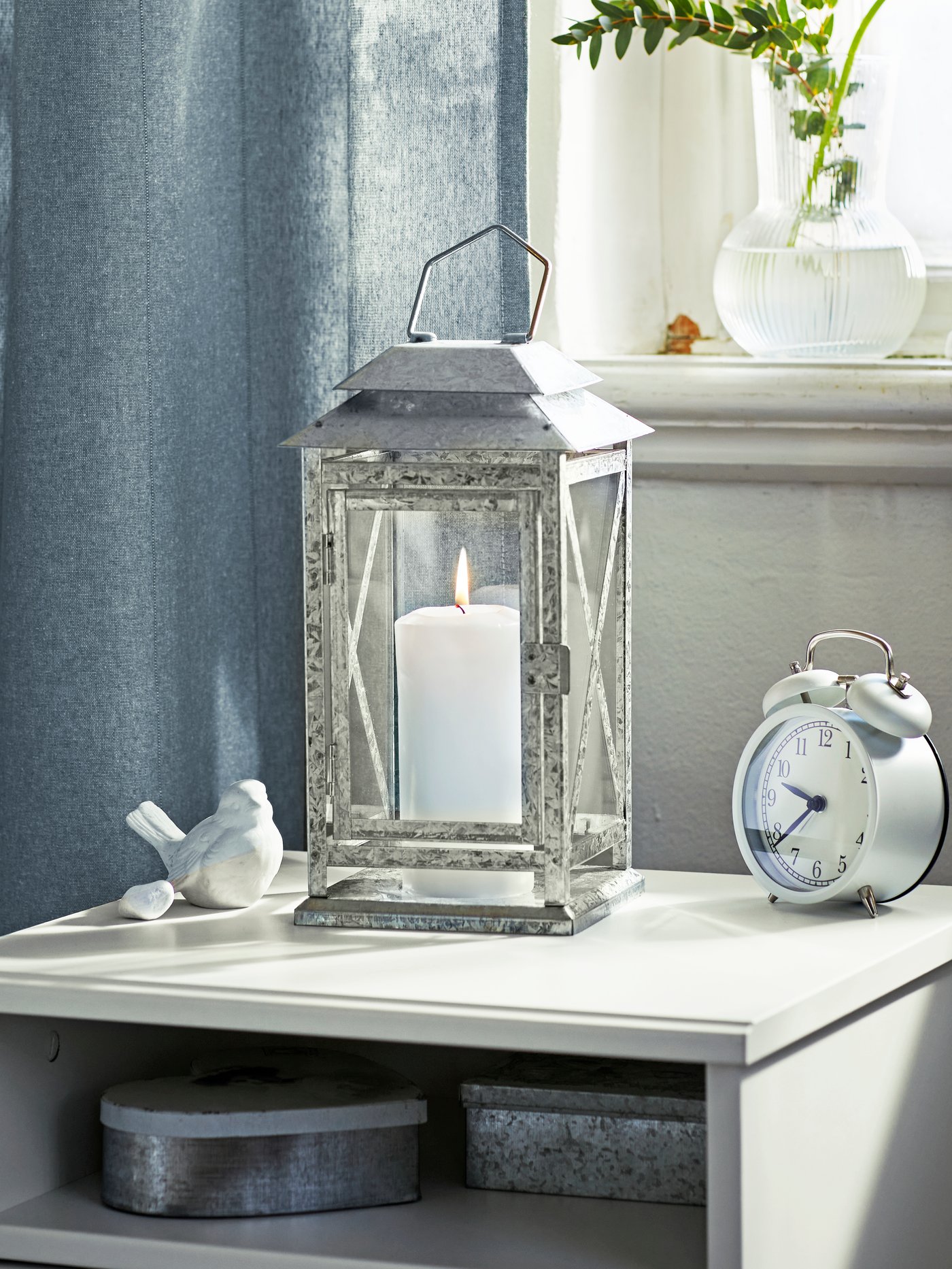 A bright, light space with a BEFÄSTA block-candle lantern and a DEKAD alarm clock placed on a SONGESAND bedside table.