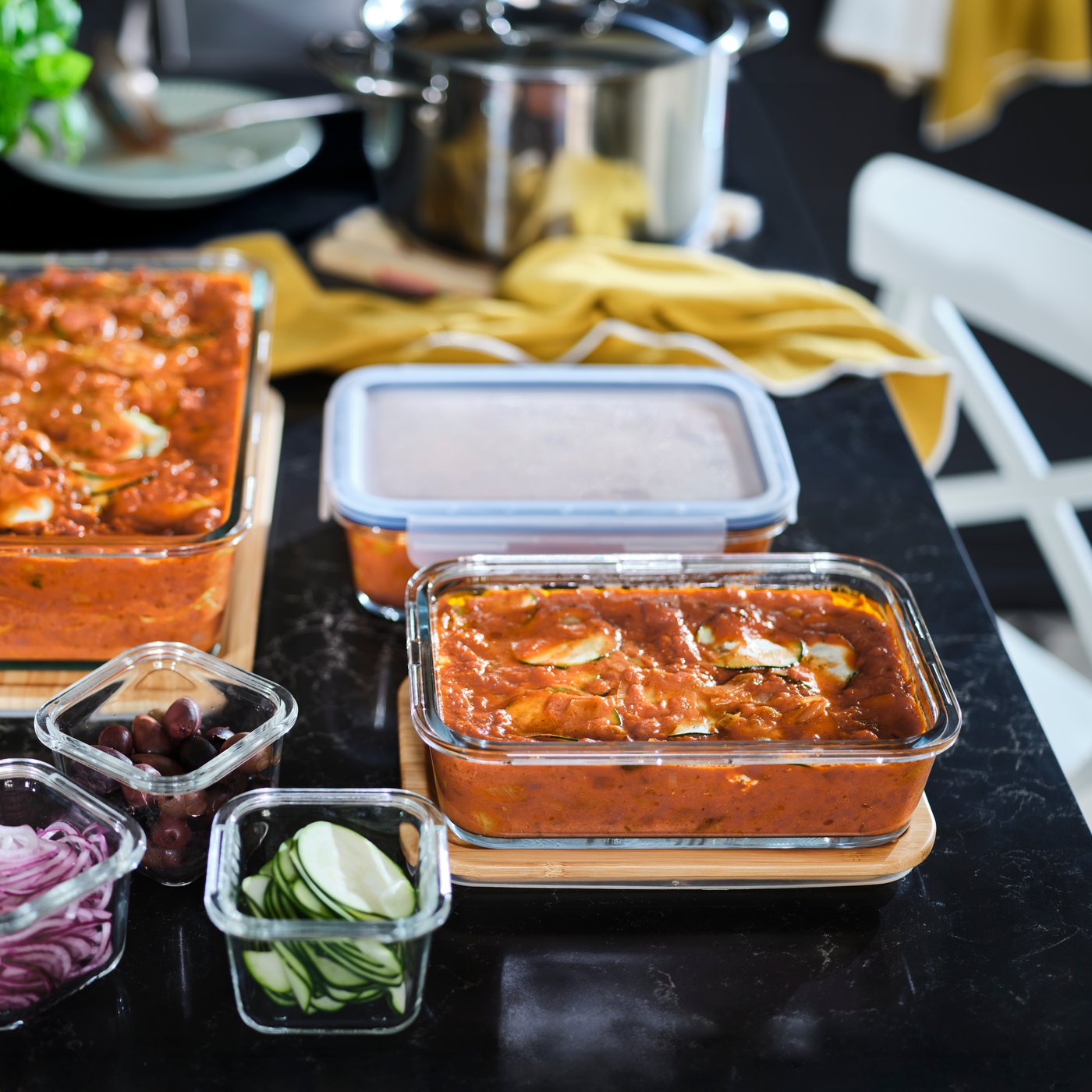 Černá kuchyňská pracovní deska, na které stojí dóza na potraviny IKEA 365+ s lasagnemi, obklopená zeleninou a dalšími jídly.