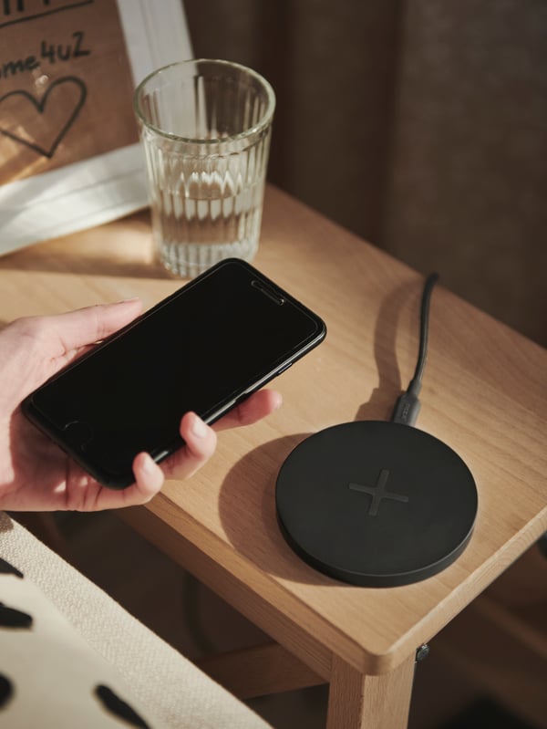 Ruka drži pametni telefon iznad crnog okruglog LIVBOJ bežičnog punjača koji se nalazi na drvenom noćnom stočiću.