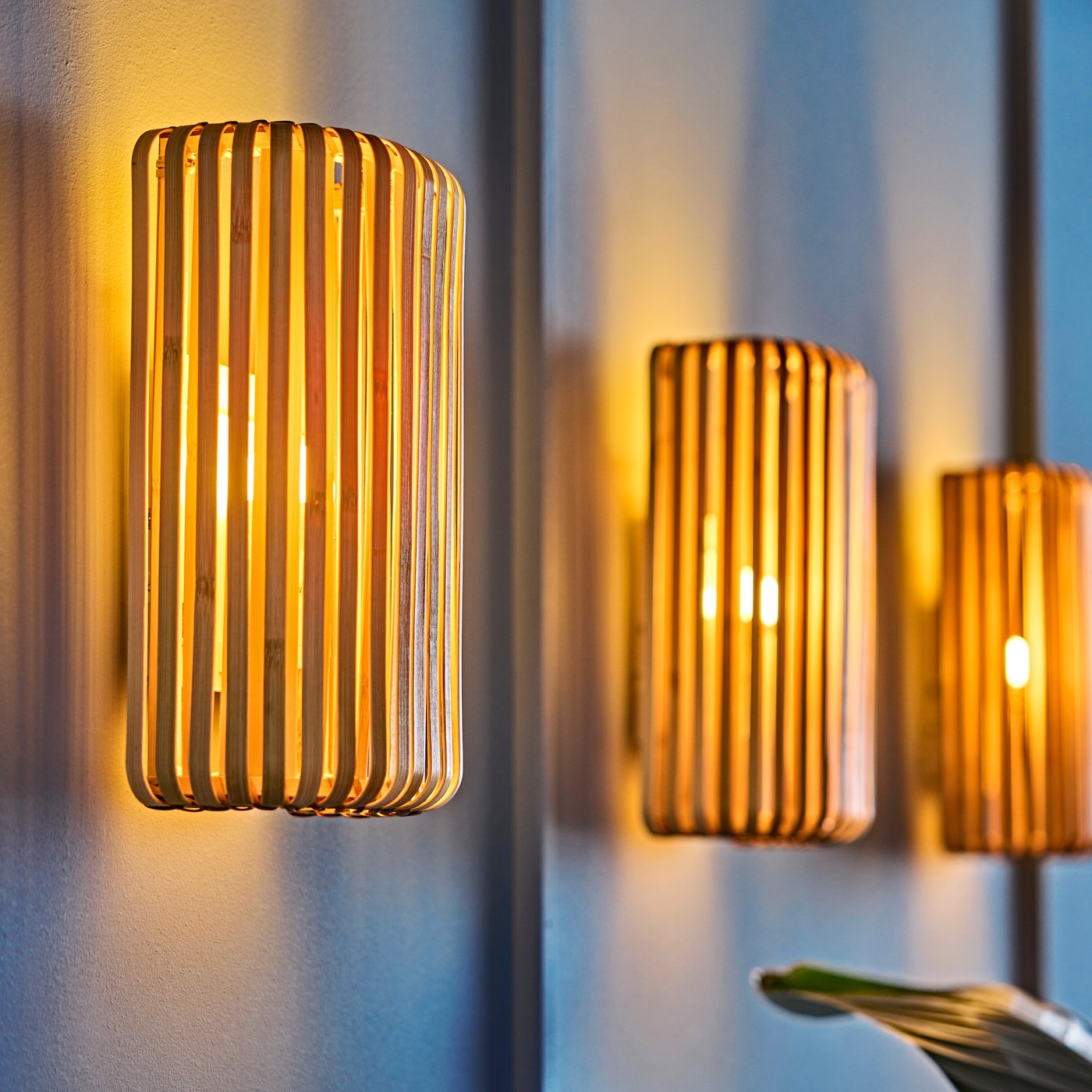 Plavi zid dnevne sobe osvetljen je toplim sjajem tri VARPTROSS zidne lampe prirodne boje napravljene od bambusa.