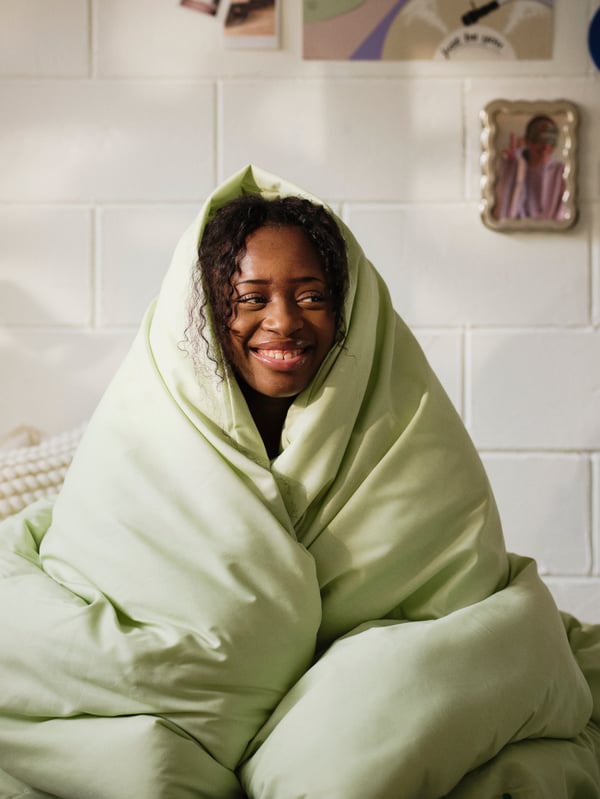 Smiling female student sitting up wrapped in light green bedding in white college dorm room.