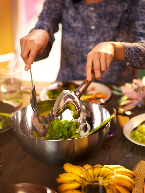 En kvinna rör om sallad i en BLANDA BLANK serveringsskål i rostfritt stål. Skålen står på ett matbord i trä tillsammans med andra serveringsfat med mat.