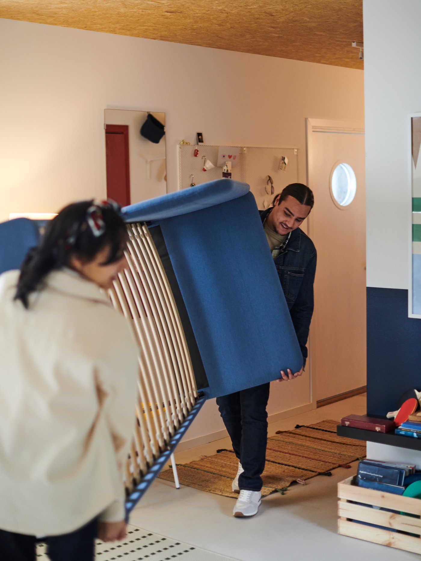 A woman and a man lifting one end each of a blue bed frame, moving it through a hallway.