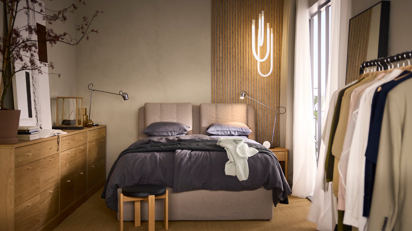 LILLFJÄLLET boxspring met opbergruimte en opgemaakt met donkergrijs bedlinnen staat tegen een beige wand met VARMBLIXT ledverlichting.