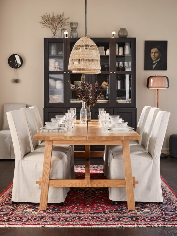 A long wooden table with glassware and traditional tableware, woven pendant lamps, chairs with beige/dark grey chair covers.