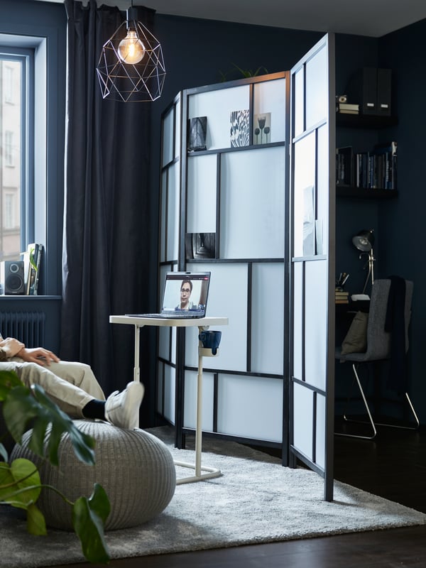 A living room divided by a RISÖR room divider, with a BJÖRKÅSEN laptop stand on one side, a compact workspace on the other.