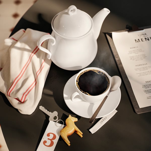 A close-up of a dining table with a white porcelain VÄRDERA coffee cup and saucer, a numbered room key, a menu and a teapot.