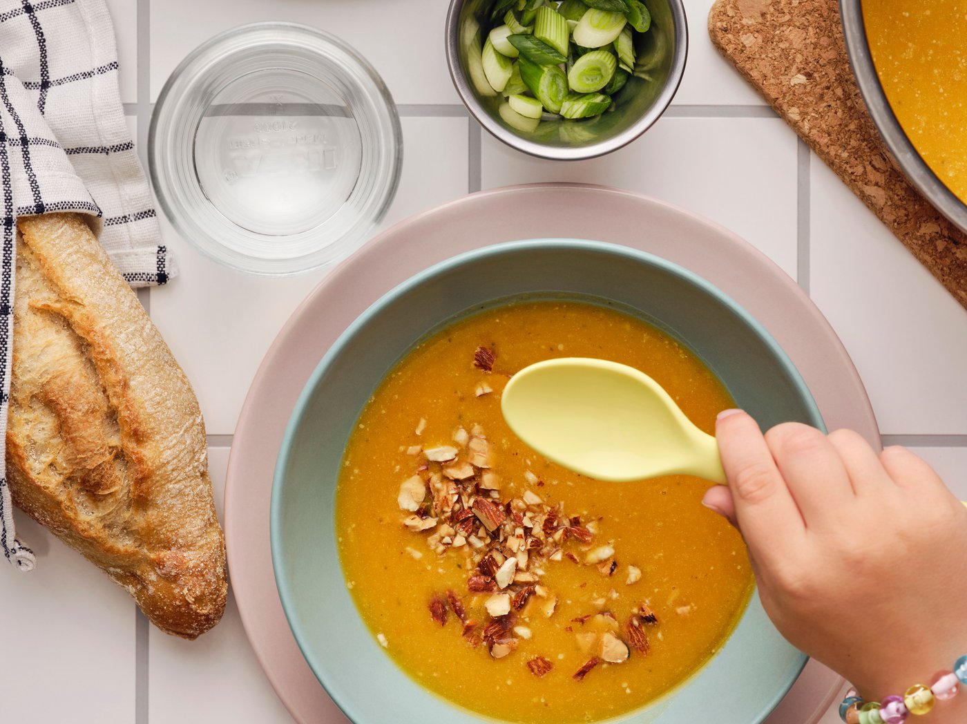 Main tenant une cuillère au-dessus d’un bol de soupe au potiron garnie d’amandes grillées MUNSBIT entouré de pain et d’aromates.