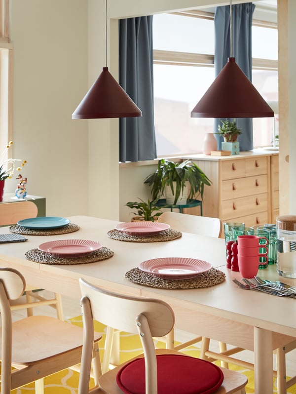 A table and chairs in birch, a yellow rug, plates in pink and blue, green glasses, cutlery and two dark red pendant lamps.