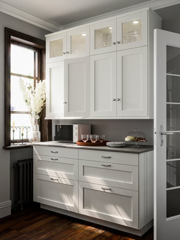 Kitchen cabinets with ENKÖPING doors and cover panels in white wood effect by a window, a microwave and items on the countertop.