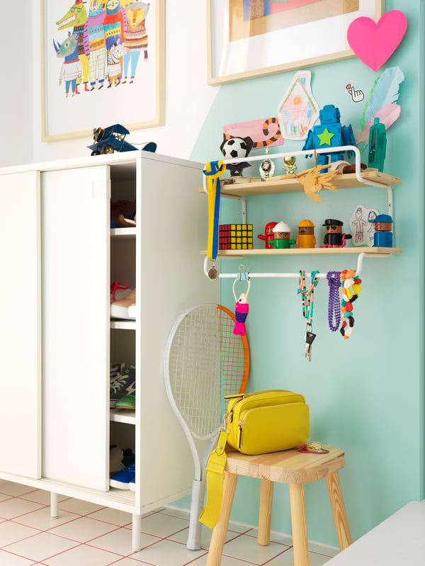 A SPORTSLIG wall shelf for trophies is attached to the wall above a FLISAT children’s stool beside a shoe cabinet.