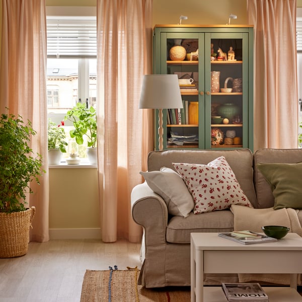 Light pink LENDA curtains hang in two windows on either side of a HEMNES glass-door cabinet behind a sofa.