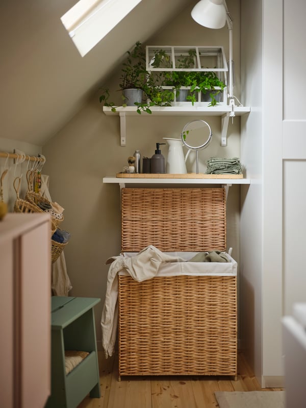 A willow NATTGIBBA laundry basket with the lid open underneath two shelves, one of which has an ÅKERBÄR greenhouse on it.