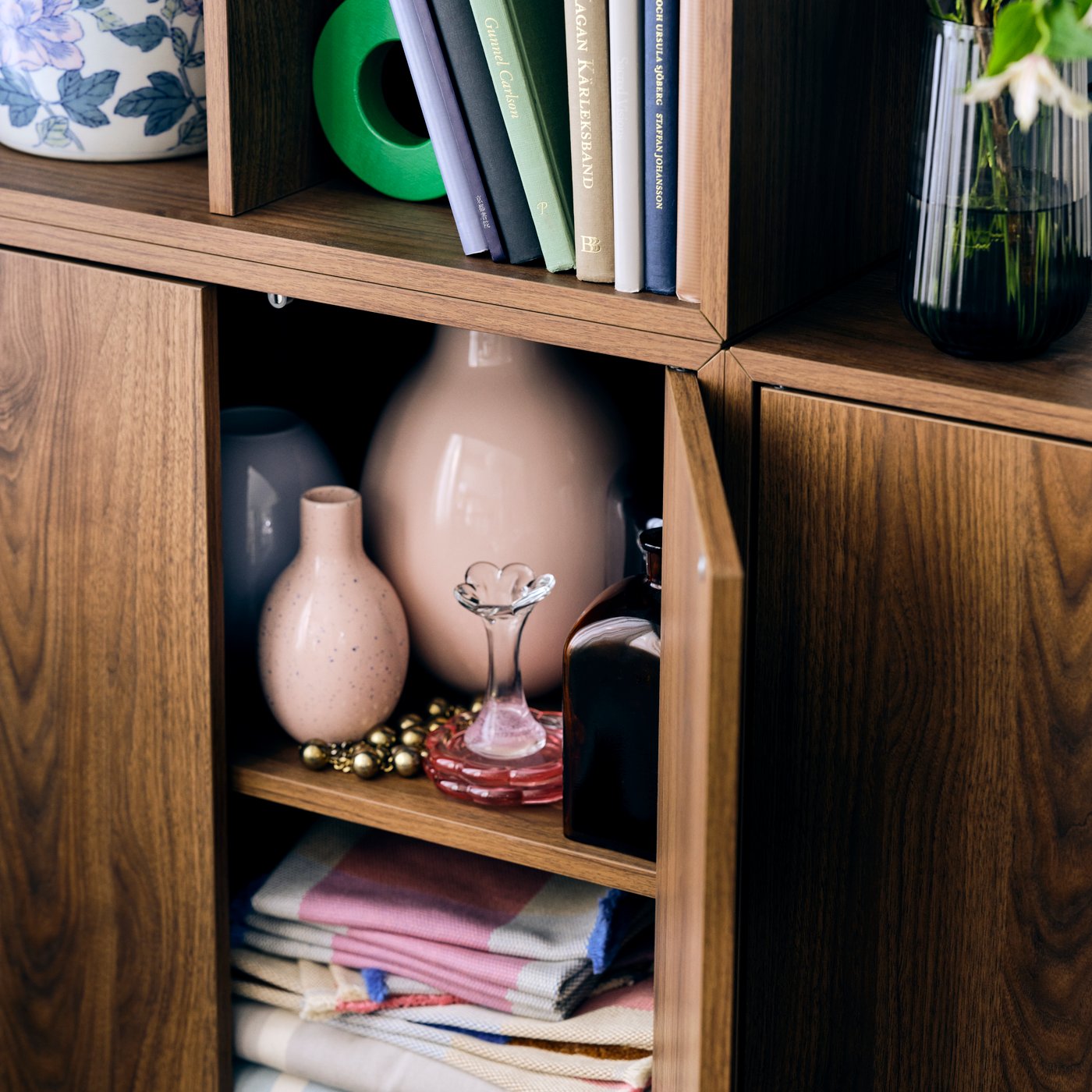 An EKET cabinet unit with the door opened to display diverse flower vases and table linen, stowed away for use on the table.