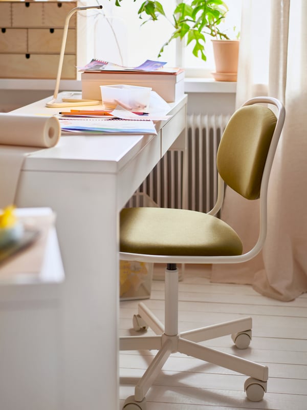 A white/yellow-green ÖRFJÄLL swivel chair, a children’s desk with a table lamp, drawings, pens and a roll of paper on top.