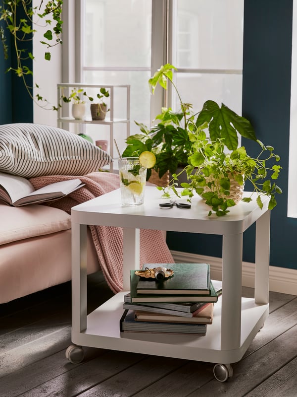 Plants on a white coffee table with wheels with a number of books on the lower level, beside a sofa and in front of a window.