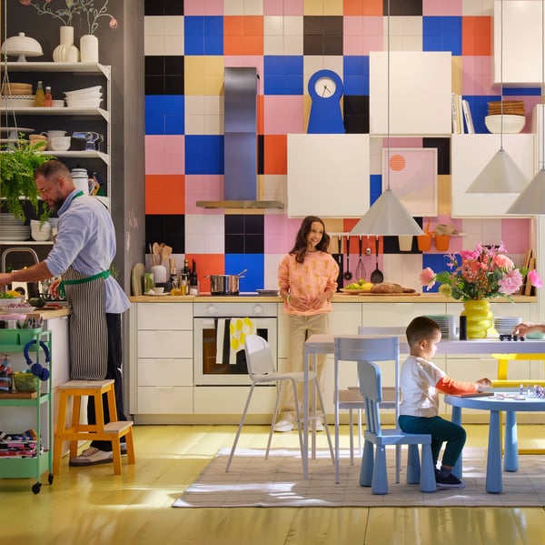 A family of three spending time together in a modern white kitchen with a high ceiling and large, brightly covered tiles.