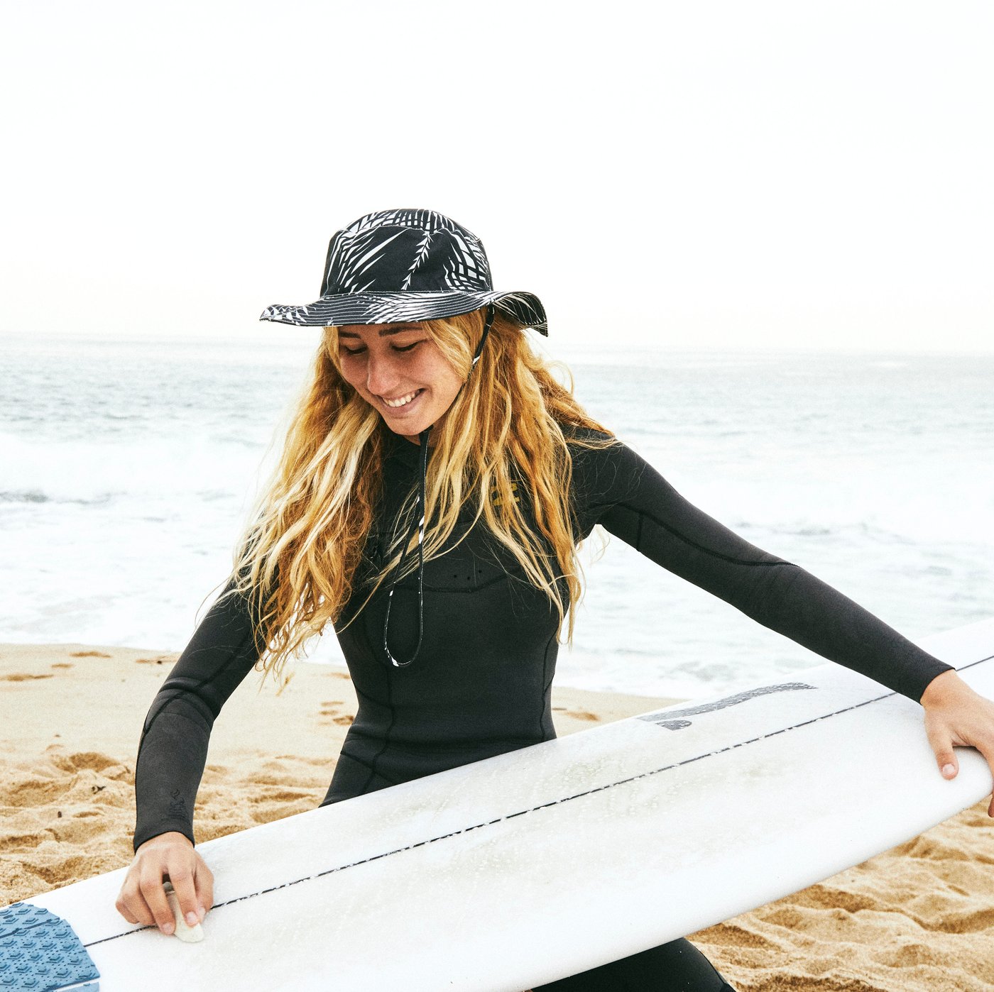 A woman wearing KÅSEBERGA hat made in a black and white palm-inspired print while holding a surfboard on a beach.