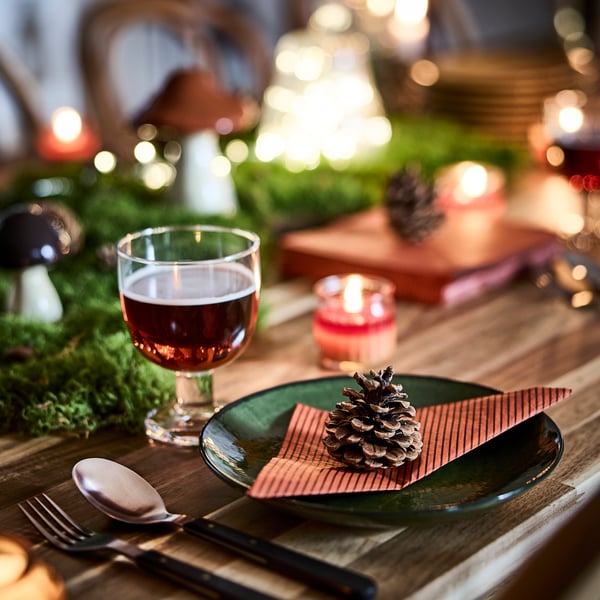 An autumn-themed table setting with a pinecone resting on a folded HÖSTAGILLE paper napkin placed on a green glass plate.