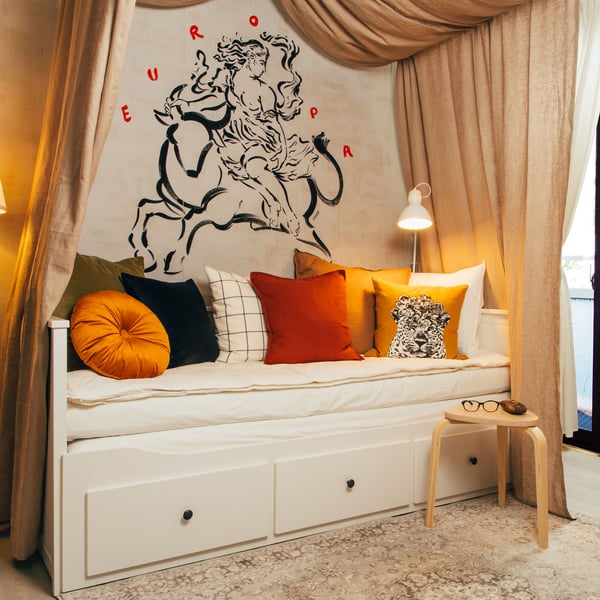 Beli sofa ležaj s mnogo jastučića u različitim bojama na vrhu. Na belom zidu je veliki crni crtež, a zavesa visi u obliku šatora.
