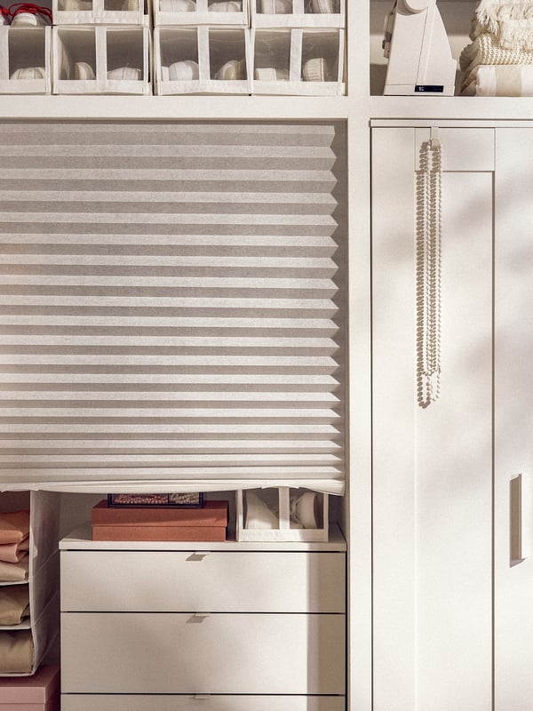 A white SCHOTTIS pleated blind attached to a high shelf beside a BRIMNES wardrobe, and pulled down in front of mixed storage.