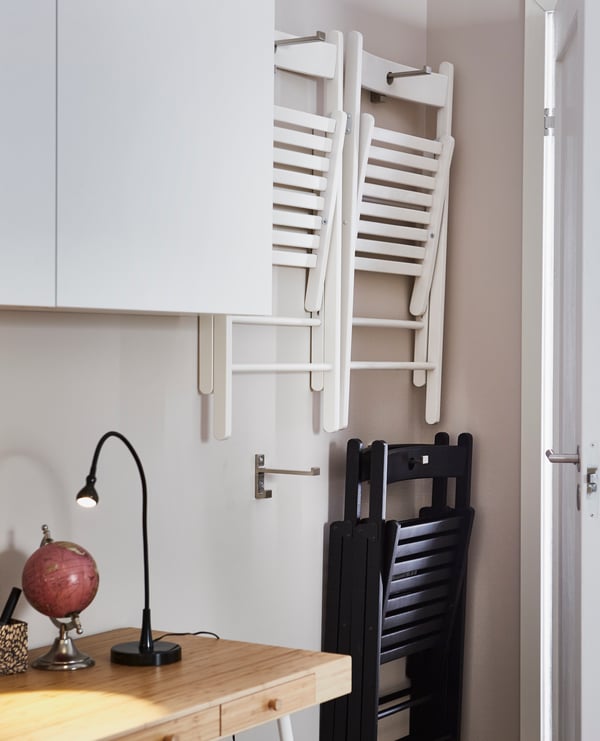 Sillas plegables colgadas en la pared de una cocina. Dos blancas arriba y una negra debajo.