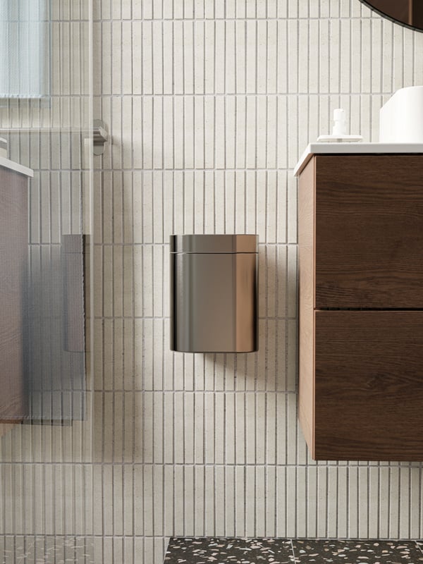 A metal trash bin is attached to a beige tiled wall in between the washbasin and the shower area.