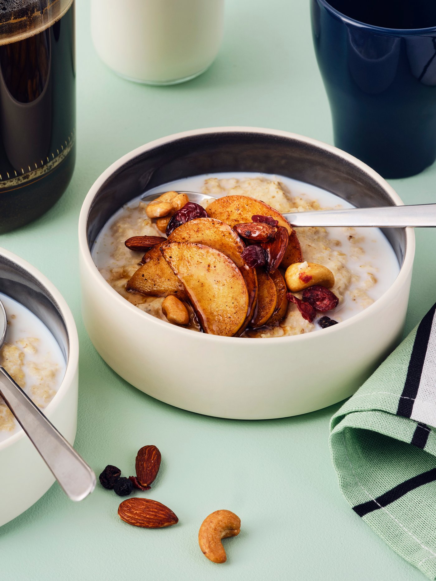 Dos tazones de avena con manzanas salteadas en canela y mezcla MUNSBIT de nueces/frutos rojos tostados, con café y leche de fondo.