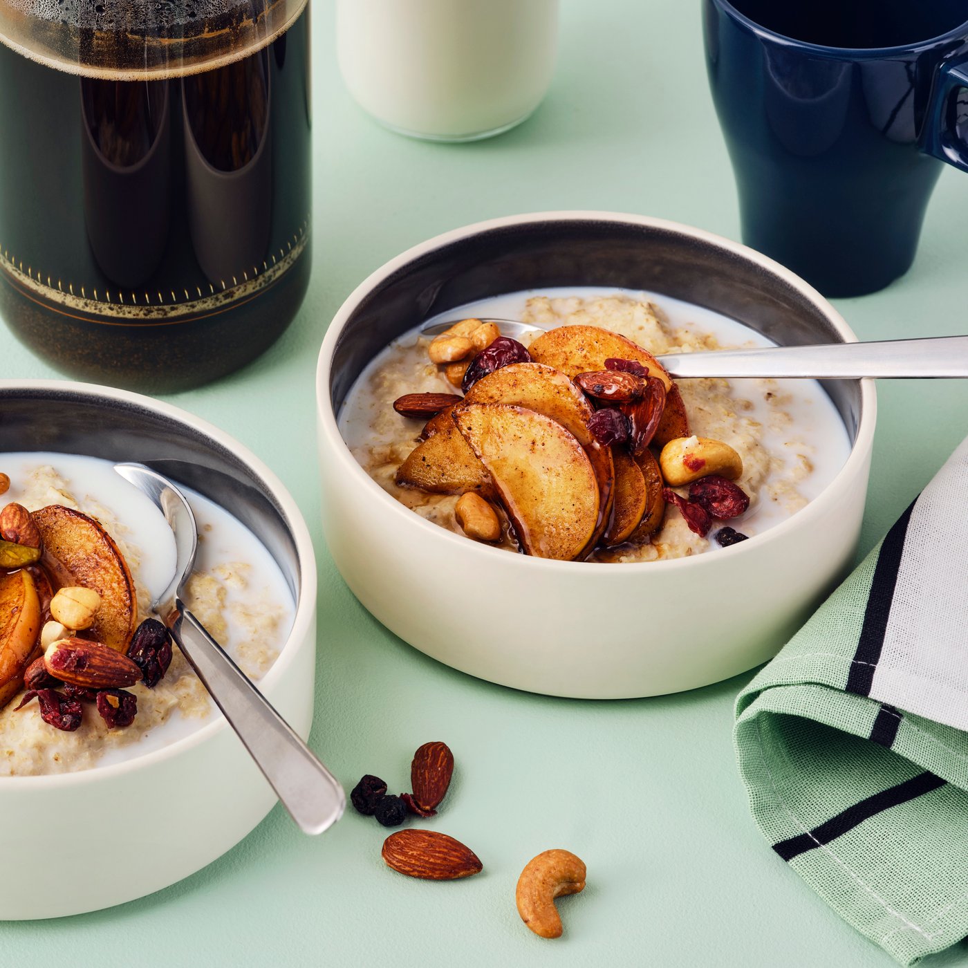 Due ciotole di porridge con mele alla cannella fritte e il mix di noci tostate/bacche MUNSBIT. Sullo sfondo, caffè e latte.