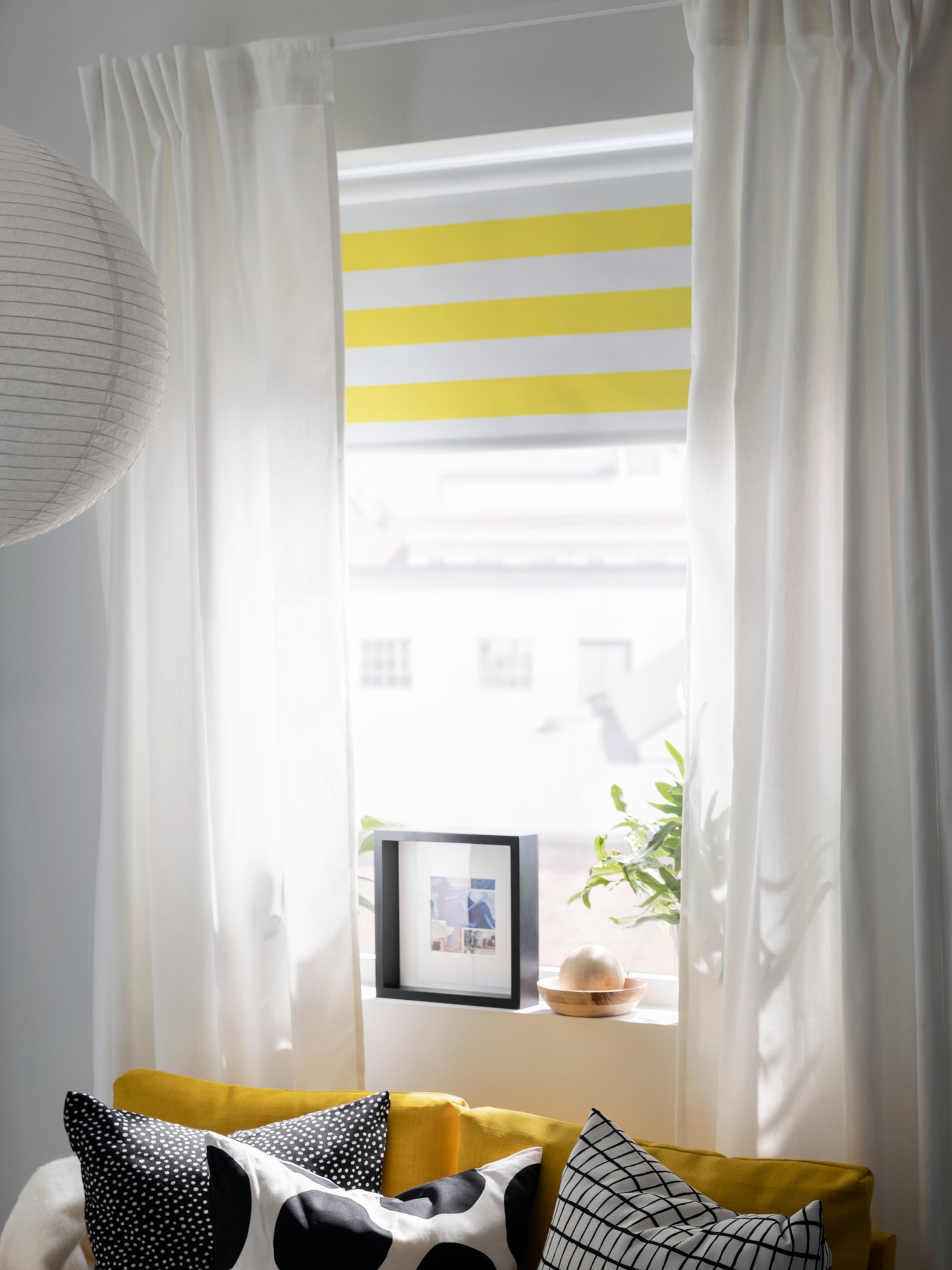 A striped FRIDANS block-out roller blind and GINSTMOTT curtains hang in a window above a yellow FRIDHULT sofa-bed.