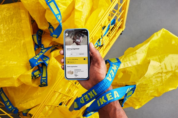 Eine Person hält ein Smartphone in der Hand, auf dem die IKEA App zu sehen ist. Um sie herum befinden sich gelbe IKEA Taschen mit blauen Griffen in einem Einkaufswagen.