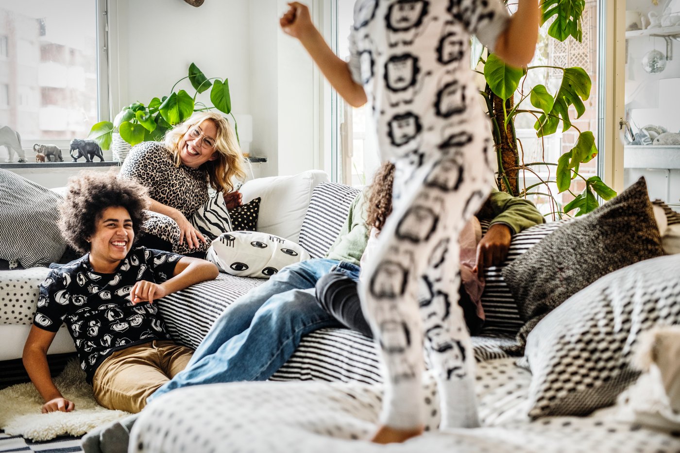 En dreng og kvinde sidder i en sofa og grinermens en tredje dreng hopper iført pyjamas i sofaen.