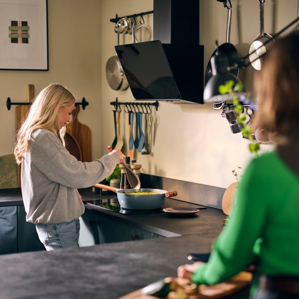 Dans une cuisine équipée d’une hotte STACKBO à puissance réglable noire, deux femmes dont l’une touille le contenu d’une casserole posée sur la plaque de cuisson.