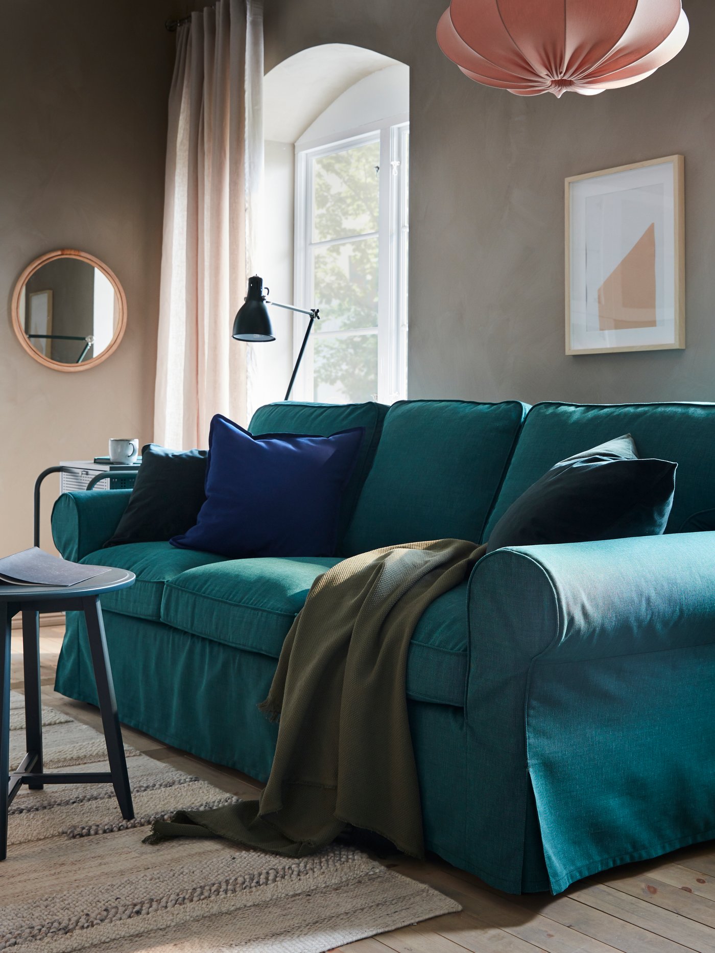 Un canapé turquoise foncé, une petite table bleu-vert foncé, un plaid vert, un tapis rayé et une suspension rose.