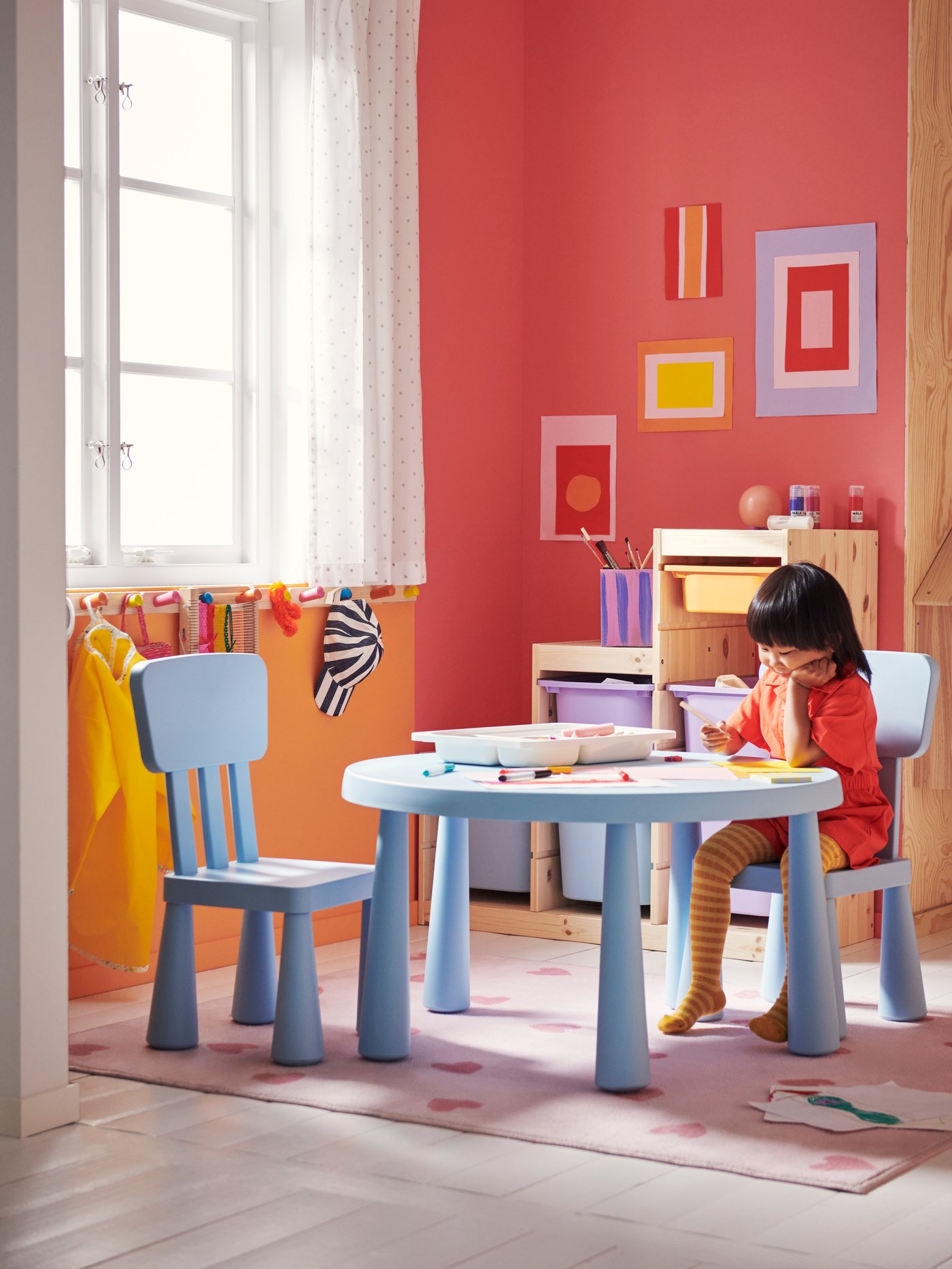 A child sitting on a MAMMUT chair at a MAMMUT children’s table drawing a picture. A tray of art materials is on the table.