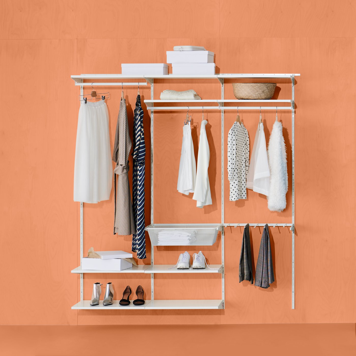 Open wardrobe against orange wall with dresses, blouses, coats, shoes, boxes and baskets on the shelves.