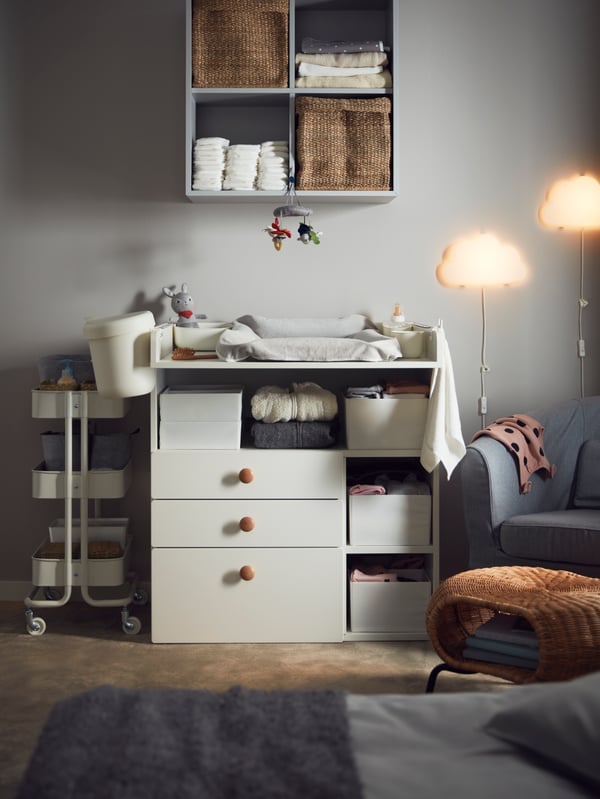A white SMÅSTAD changing table with 3 drawers, a natural material footstool, a storage trolley and a grey armchair in a cosy room.